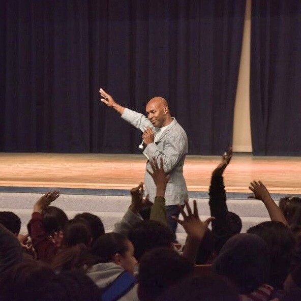 Ken Shelton speaks to an auditorium of excited students waving their hands in the air.